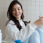 Smiling woman holding a glass while seated, with pill bottles on the table.