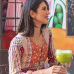 Woman holding a refreshing green juice in a lively café setting with colorful decor.